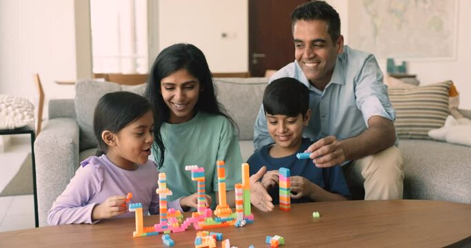 Happy Indian spouses play build blocks with little kids. Siblings sit on floor at table in cozy living room enjoy playtime with loving young pretty mom and dad. Family weekend leisure at home, games