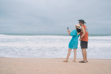 Asian tourist couple taking selfies, sand on beach and blue summer sky with white waves, summer vacation and holiday concept.