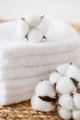 stack of white towels on woven mat and branch of cotton with white cotton bolls. white textured wall. Concept of luxury, relaxation, self-care, home decor, spa, and wellness.