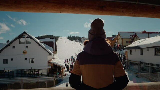 View from the hotel balcony to the snowing ski path resort. Back view of woman dancing while standing on winter resort balcony looking the tourists.