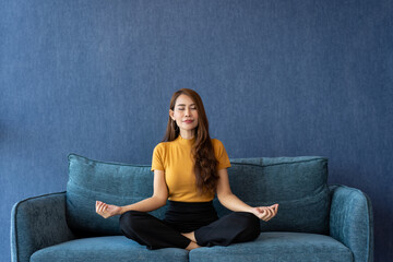Portrait of healthy Asian woman meditating and exercise pose yoga on the sofa in morning. Zen...