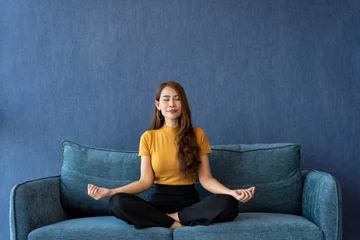 Fotobehang Portrait of healthy Asian woman meditating and exercise pose yoga on the sofa in morning. Zen relaxation and mindfulness lifestyle, Concept of indoor workout for good healthy and breathing practice. © Prot