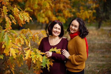 Warm autumn mood. Beautiful middle-aged woman with her adult daughter are smiling and walking in park on background of beautiful autumn leaves. Fall coming. Autumn family story. Yellow leaves in park