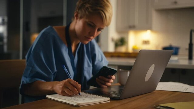 Tired woman wearing medical scrubs looking at mobile phone whilst working or studying on laptop at home at night - shot in slow motion