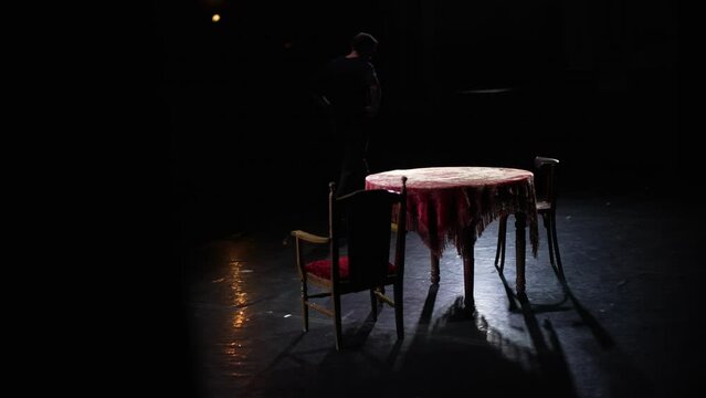 The actor walks across the stage next to an old table and chairs.
