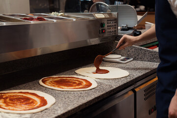 Anonymous male chef spreading tomato sauce with big iron spoon onto pizza while preparing pizza in...