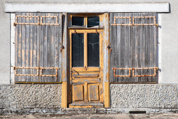 Old weathered, half-decayed and broken yellow painted wooden door with side windows with old wooden shutters in front, in a dilapidated house with a plastered white wall in France