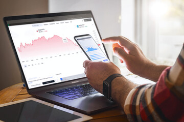 Investor trader analyzing financial trading crypto stock market. Man using mobile phone and laptop checking and doing investing analysis, working with charts and market reports