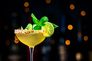 Spicy cucumber margarita cocktail drink with tequila, lime juice, syrup, mint and ice in glass garnished with salted pepper rim, dark background, bar tools and bottles