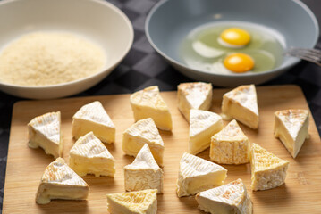 Sliced Camembert cheese on a wooden cutting board against a background of browned plates with breadcrumbs and chicken eggs