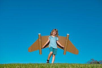 Happy child with paper wings against blue sky. Kid with toy jetpack having fun in spring green field outdoor. Freedom carefree and kids imagination dream concept.