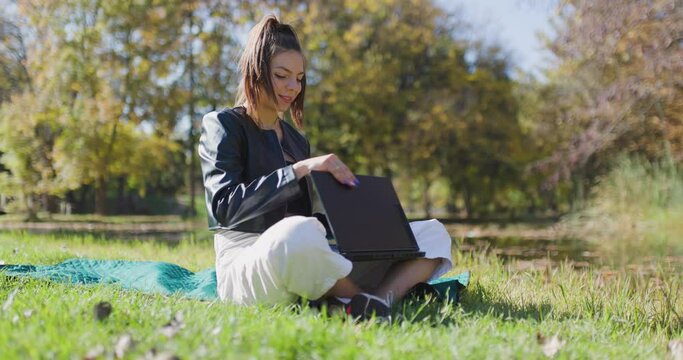 Beautiful girl working on a laptop computer and lying on a meadow. Fashionable young lady in the park. Left camera movement.