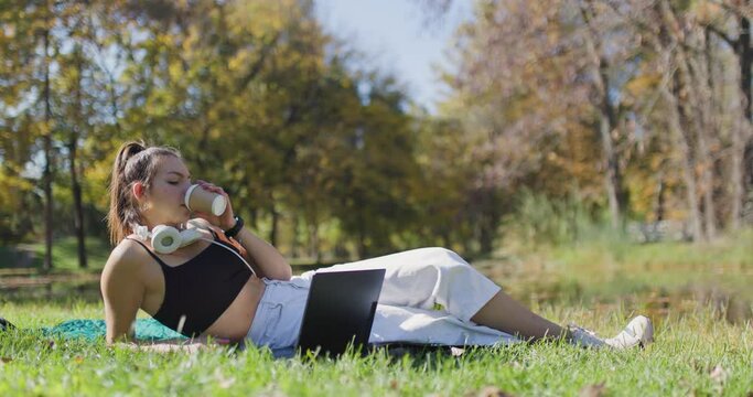 Beautiful girl with laptop and headphones drinking coffee from a recyclable coffee cup. Fashionable young lady lying in the park.