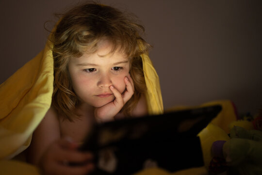 Kid playing game on tablet in bed at night. Kids with social media. Child lying in bed playing a tablet in dark room, light under blacket. Close up of kid watching cartoons on tablet.
