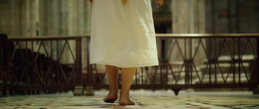 Barefoot tramp woman wanderer looking for God in church. Parishioner holding a religious cross in hand and walks along the pews.