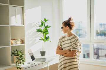 Young adult woman standing next to a work desk with a laptop in a modern apartment near the window. Home office concept
