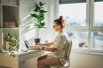 Young adult Caucasian woman in glasses working on a laptop in a modern apartment office near the window