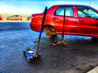 A red car with a punctured wheel and a jack for its repair on the asphalt outdoors on a city street. Car repair service. Roadside assistance