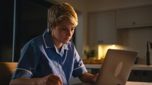 Tired woman wearing nurse's uniform working or studying on laptop at home at night drinking hot drink - shot in slow motion
