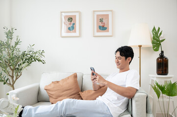 Happy asian man sitting on couch using smartphone in living room at home, relax time and lifestyle concept