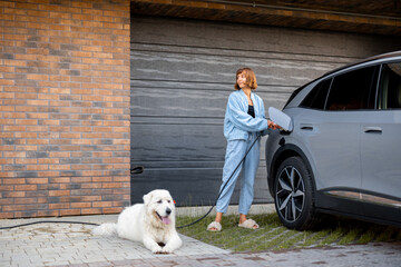 Young woman charges her electric car while standing with her cute dog near garage of her house. Concept of modern and happy lifestyle