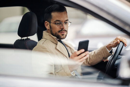 A young elegant businessman is leaning on a pillar at station and waiting for transport while using his phone.