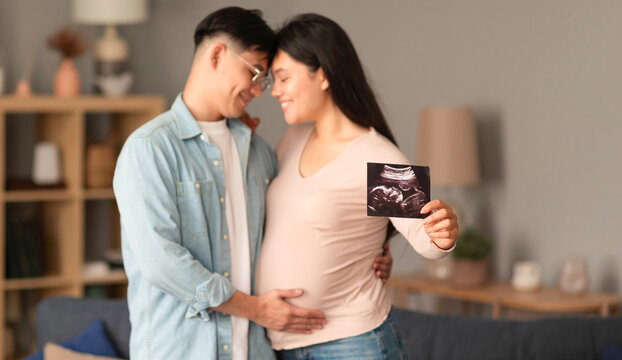 Japanese couple embracing, pregnant woman showing ultrasound photo at home