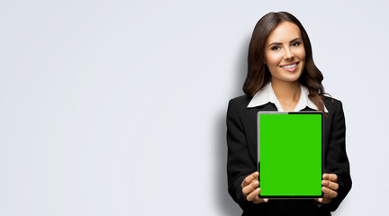Fototapeta premium Portrait of happy smiling brunette businesswoman showing tablet pc, touchpad, with green chroma key screen, against lightly grey gray background. Confident attractive business woman at office.