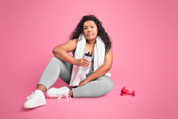 Tired black body positive woman in sportswear sitting on floor, holding bottle of water, have break...
