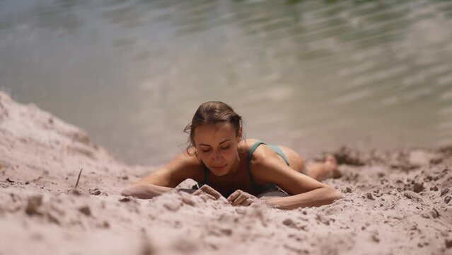 tanned young slim woman with sporty body lying outdoors on hot sand on lake shore and sunbathing. refreshing in heat, natural outdoor lifestyle. relaxing in nature