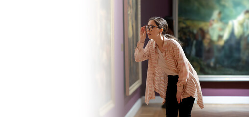 Fototapeta premium Web banner of maseum day. Side view of young pretty Caucasian woman carefully looking at picture. Masterpieces in background. Mock up