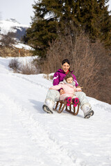 Front view of a happy mother and daughter riding wooden sled down snowy hill enjoying a family day out. Spending time together outdoors on a sunny day with the mountain in the background. Xmas days.