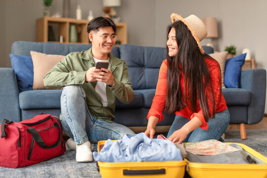 Asian man using smartphone booking vacation, woman packing suitcase indoor - Powered by Adobe