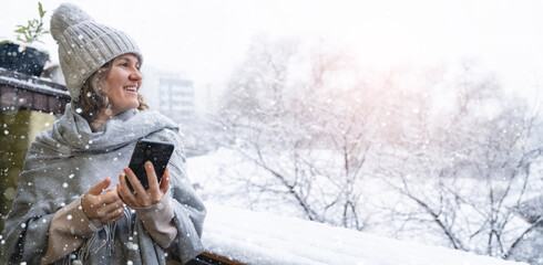 A woman in a warm hat and blanket holds smartphone in her hands. Winter landscape with snowfall in...