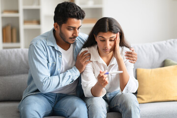 Unwanted pregnancy. Worried hindu couple looking at positive test result, sitting on sofa at home - obrazy, fototapety, plakaty