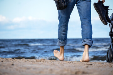 On the seashore of blue color, a man in denim pants walks on the beach sand barefoot and with black...