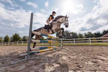 Girl rider on dapple gray horse jumping over triple bars in outdoor arena on a sunny summer day....