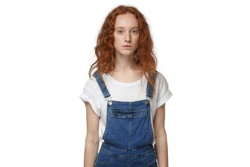 Fototapeta premium Young redhead woman standing still in denim jumpsuit, looking at camera