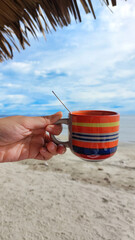 Holding up a cup of coffee served in a colorful boho styled cup at the sunny beach, relaxing and unwinding on a summer day