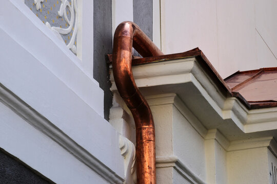 shiny new copper rain water leader, downspout or down pipe detail. lightning protection grounding rod. home renovation, construction concept. textured white stucco exterior wall and window background