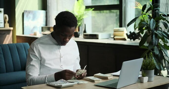 A diligent Black businessman sits at desk, meticulously counting cash with the assistance of a calculator. His focused approach to financial matters is evident in this scene of financial management.