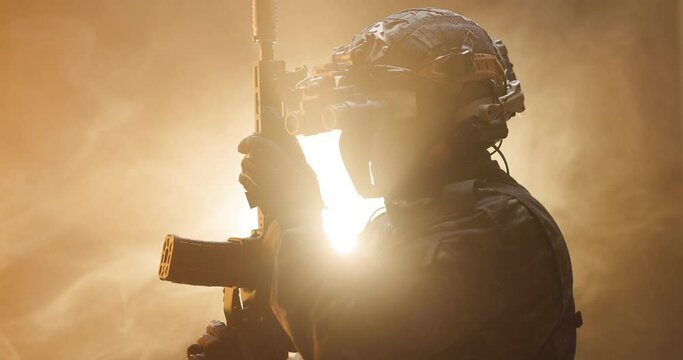 Soldier fully equipped with tactical gear and protection aiming his rifle at enemy with orange smoke in the background.