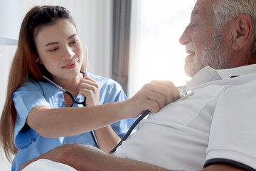 Obraz premium Smiling beautiful caring female doctor using stethoscope listen to senior patient heart who lying in hospital bed. Nurse takes care man feel sick and rest in bed at home, medical elderly health care.