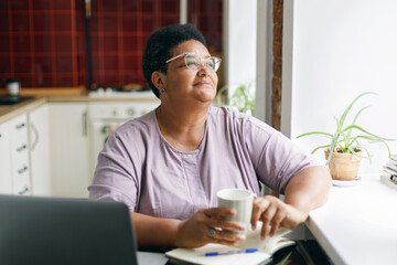 ensive thoughtful senior plus size woman of african ethnicity in spectacles sitting at kitchen table with coffee in hands looking through window contemplating, having rest after working on laptop