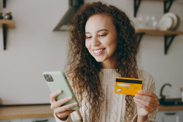 Young happy housewife woman wear casual clothes sweater use mobile cell phone credit bank card shopping online buy products sit at table in light kitchen at home alone. Lifestyle cooking food concept.