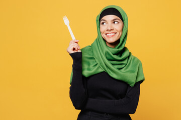 Young smiling happy arabian asian muslim woman wear green hijab abaya black clothes hold fork look aside on area isolated on plain yellow background. People uae middle eastern islam religious concept.