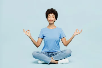 Poster Im Rahmen Full body young woman of African American ethnicity in t-shirt casual clothes sit hold spread hand in yoga om aum gesture relax meditate try to calm down isolated on plain light blue cyan background © ViDi Studio