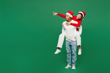 Full body young couple man woman in red casual clothes Santa hat posing give piggyback ride to...