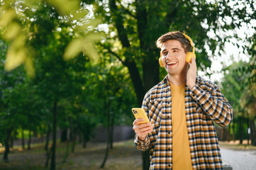 Young man wear casual clothes headphones listen music use mobile cell phone walk rest relax in spring green city park go down alley sunshine lawn outdoors on nature. Urban lifestyle leisure concept.