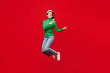 Full body young woman wear green turtleneck Santa hat posing jump high point index finger aside on mock up isolated on plain red background. Happy New Year 2024 celebration Christmas holiday concept.